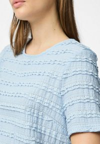 Gaiši zilas krāsas topiņš, kas izgatavots no teksturēta auduma ar kārtām un rūtotiem svītrām, kā arī ar smalkiem izgriezumiem. Apaļš kakla izgriezums un īsās piedurknes.