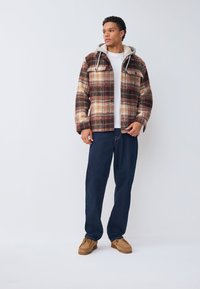 Chaqueta de cuadros en tonos marrón, crema y óxido con capucha, bolsillos frontales y cierre de botones; combinada con pantalones azul oscuro y zapatos marrones.