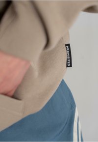 Beige genser med ribbet tekstur, med en sidelomme. Blå bukser under med subtile sømmer og en hvit vertikal stripe detalj.
