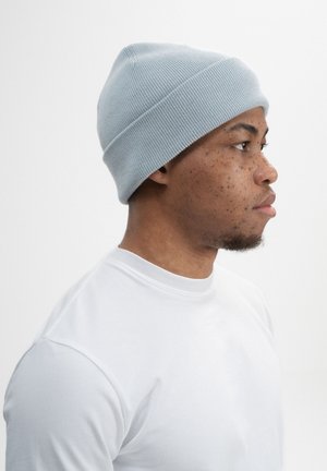 Junger Mann im Profil, der eine hellblaue gestrickte Mütze und ein weißes T-Shirt mit Rundhalsausschnitt vor weißem Hintergrund trägt.