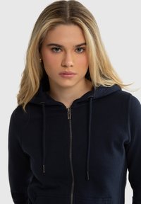 Blonda sieviete ar taisniem matiem valkā tumši zilu rāvējslēdzēja kapuci ar auklām, skatoties taisni uz priekšu pret vienkrāsainu fonu.