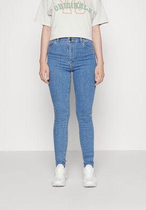Personne portant un jean skinny bleu clair, des baskets blanches et un t-shirt court blanc avec un texte vert et orange.