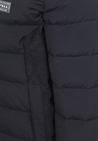 Donsjack in donker marineblauwe stof met horizontaal doorgestikte secties, voorzien van een ribbeltextuur zijpaneel en merkpatchdetail.