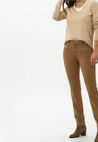 Beiger V-Ausschnitt-Pullover kombiniert mit braunen, figurbetonten Hosen mit Reißverschluss-Vordertaschen. Das Outfit umfasst braune Stiefeletten.