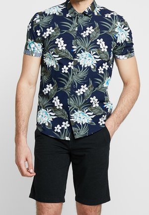 Mann trägt ein marineblaues Kurzarmhemd mit weißem und hellblauem Blumenmuster und schwarze Shorts, steht mit einer Hand in der Tasche.