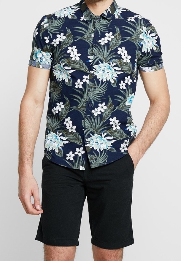 Homme portant une chemise à manches courtes bleu marine avec un imprimé floral blanc et bleu clair et un short noir, debout avec une main dans la poche.