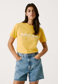 Gelbes Baumwoll-T-Shirt mit weißem "Red Bull Racing"-Logo, kombiniert mit hellblauen Jeansshorts mit fransigen Kanten.