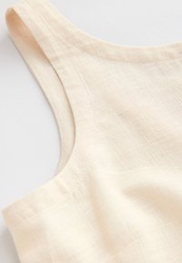 Vêtement sans manches beige clair avec un col rond, présentant un tissu texturé et des détails de coutures visibles le long des bords.