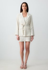 Blazer in lino beige chiaro con cintura a cravatta, abbinato a una gonna attillata coordinata. Presenta sottili righe e bottoni. Sandali argentati con cinturini.