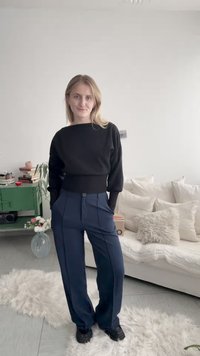 Ung kvinde med blondt hår iført sort langærmet top og marineblå bukser står i en stue med en hvid sofa og et grønt sidebord.