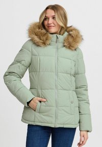 Chaqueta de plumas verde claro con capucha de pelo sintético, diseño acolchado, cremallera frontal y bolsillos laterales. Adecuada para usar en clima frío.