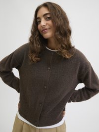 Jovem mulher com cabelo castanho ondulado, vestindo um cardigan castanho escuro com botões sobre uma camisa branca e calças beiges, sorrindo ligeiramente, com as mãos na cintura.