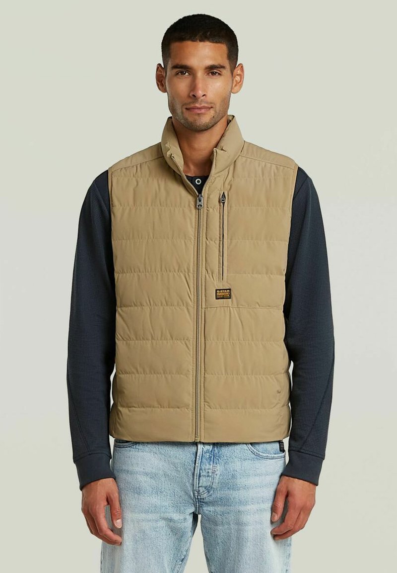Mann trägt eine beige gesteppte Weste über einem dunklen Langarmshirt und hellblauen Jeans, steht vor einem einfarbigen Hintergrund.