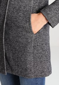 Manteau texturé gris foncé avec un motif tissé, doté de poches latérales et d'une coupe droite, porté sur un jean bleu.