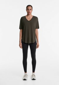 Oliven grøn V-hals t-shirt i en løs, flydende design, parret med sorte tætsiddende leggings og hvide sportssko. Glat tekstur, afslappet pasform.