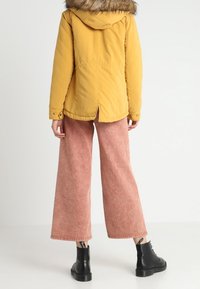 Żółta parka z kapturem wykończonym futrem, zestawiona z różowymi spodniami o szerokich nogawkach i czarnymi botkami za kostkę. Gładka faktura i casualowy fason.