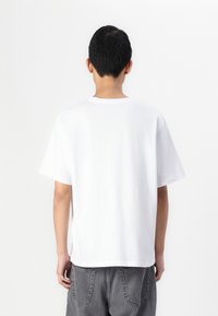 Maglietta bianca in cotone con collo rotondo, maniche corte e vestibilità rilassata, vista da dietro. Design semplice e uniforme, senza motivi.