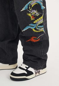 Black denim pants with vibrant embroidered dragon and waves; paired with black and white sneakers featuring a skull design on the side.