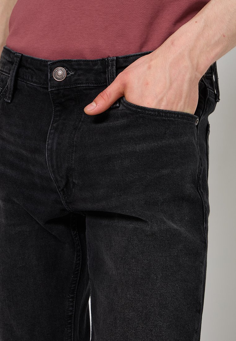Czarne dżinsy o slim fit, z pięcioma kieszeniami i zapięciem na guzik z przodu, pokazane z ręką spoczywającą w kieszeni.