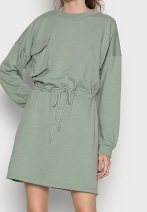 Vrouw draagt lichtgroene jurk met lange mouwen, een trekkoord in de taille en groot zakdetail op de borst, staand tegen een effen achtergrond.