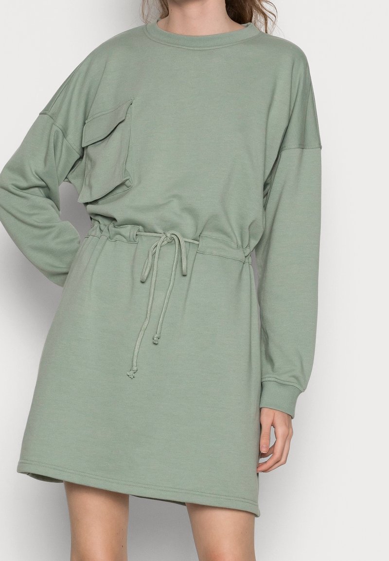Femme portant une robe à manches longues vert clair avec une taille à cordon et un grand détail de poche sur la poitrine, debout devant un fond uni.