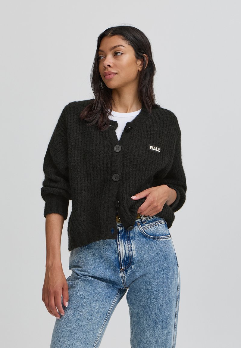 Cardigan negru cu banda, cu închidere cu nasturi, având un patch cu logo pe partea stângă. Combinat cu blugi albaștri cu talie înaltă.