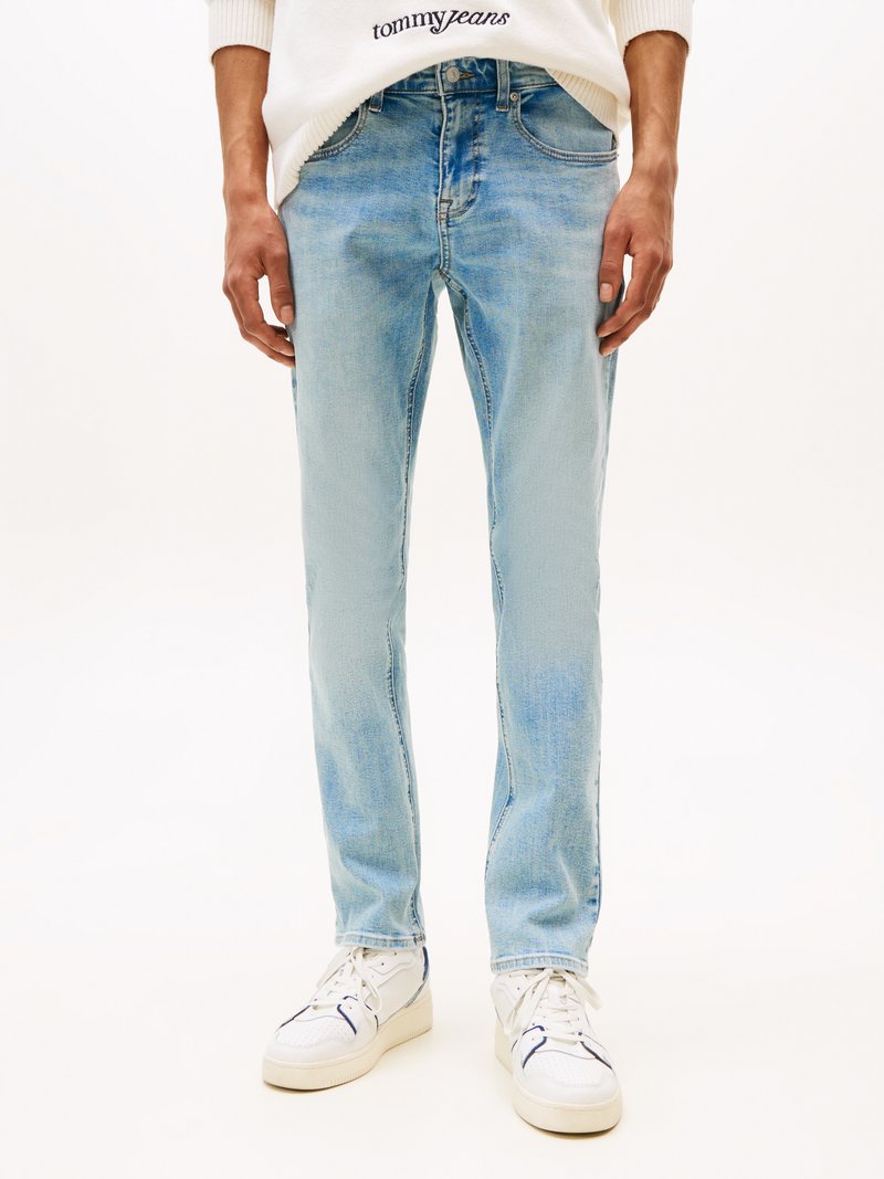 Light blue denim jeans with a slim fit, faded wash, five-pocket design, and a subtle logo on the waistband. Styled with white sneakers.