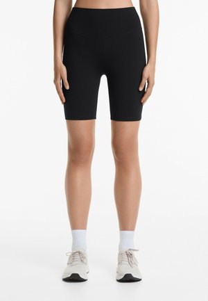 Zwarte high-waisted fietsbroeken, gemaakt van rekbaar materiaal, met een gladde textuur, knie-lange snit, gecombineerd met lichtgekleurde sneakers en witte sokken.