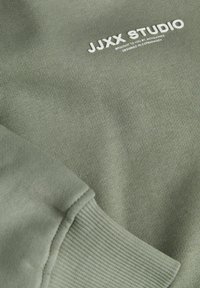 Olivově zelená látka s žebrovaným manžetovým koncem a bílým textem "JJXX STUDIO přináší vám Jack & Jones, navrženo v Kodani."