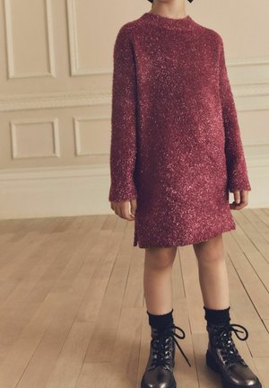 Robe tricotée brillante couleur bordeaux avec manches longues, col rond et fentes latérales ; associée à des bottines noires à lacets.