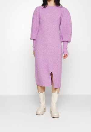 Kobieta ubrana w lawendową, dzianinową sukienkę midi z bufkami na długich rękawach i rozcięciem z przodu, połączoną z kremowobiałymi, masywnymi kozakami do połowy łydki.