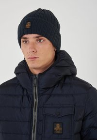Cappello in maglia a coste di colore blu scuro con patch del logo, indossato con un giubbotto imbottito con colletto alto e chiusura a zip, e una tasca sul petto.