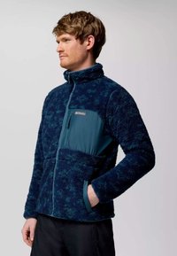 Giacca in pile di colore verde scuro, con chiusura a zip, colletto alto e pannello liscio a contrasto. Include tasche frontali in tessuto testurizzato.