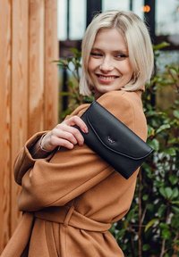 Černá kožená peněženka s hladkým povrchem, zakřiveným klopou a decentně embosovaným logem. Modelka má na sobě béžový kabát a drží peněženku u ramene.