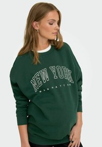 Sudadera verde de corte holgado, con escote redondo y puños acanalados, que presenta "NUEVA YORK" y "MANHATTAN" impresos en blanco.