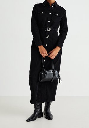 Vrouw draagt een lange zwarte jurk met knopen en een riem, houdt een kleine zwarte handtas vast, en draagt zwarte leren laarzen tot halverwege de kuit, tegen een effen achtergrond.