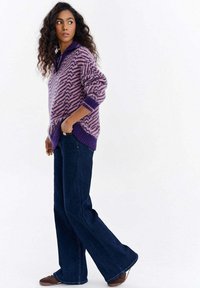 Sweter dziany w różowo-fioletowy wzór jodełki z wysokim kołnierzem, zestawiony z ciemnoniebieskimi rozszerzanymi dżinsami i brązowymi butami.