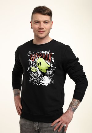 Sorte sweatshirts med et grafisk print, der viser et grønt spøgelse og stiliseret tekst. Ærmerne og kanten er ribbet for et tætsiddende look.