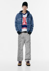 Felpa blu con testo, sovrapposta a una maglietta bianca e rossa a righe. Pantaloni grigi pixelati e scarpe marroni con lacci blu completano l'outfit.