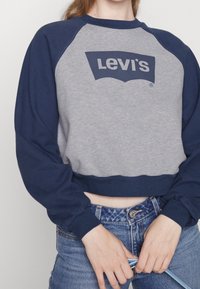 Sudadera recortada en gris y azul marino con mangas largas, que presenta un gran logotipo negro de "Levi's" en el frontal, fabricada en suave material de algodón y con ribete acanalado.
