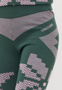 Grønne leggings med et geometrisk rosa mønster, vertikale striper og en teksturert finish. Merkevarenavn "KARI TRAA" er diskret plassert på livet.