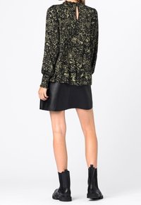 Femme portant un blouse à manches longues noire et verte à motif, une mini-jupe en cuir noire et des bottes noires à la cheville, se tenant de dos.