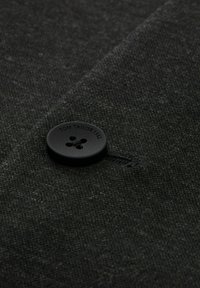 Ciemnoszary materiał o teksturowanym splocie, z czarnym okrągłym guzikiem z wygrawerowanym napisem "TOM TAILOR 1962" oraz czarną nitką.