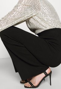 Pantaloni neri a gamba larga abbinati a una blusa chiara con decorazioni di paillettes, che mette in risalto una vita aderente, completati da sandali neri con tacco alto.
