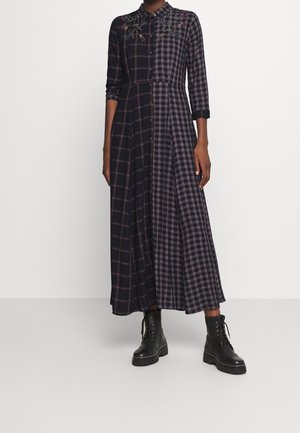 Mujer que lleva un vestido maxi de manga larga y abotonado con patrones de cuadros mezclados y bordado floral en la parte superior, combinado con botas negras con cordones.