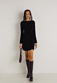 Abito mini nero a maniche lunghe con un design aderente e dettagli di arricciatura sottili. Abbinato a stivali alti fino al ginocchio in pelle marrone e a una borsa coordinata.