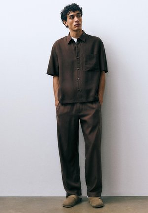 Jeune homme aux cheveux bouclés portant une chemise marron à manches courtes boutonnée et un pantalon assorti, debout les mains dans les poches.