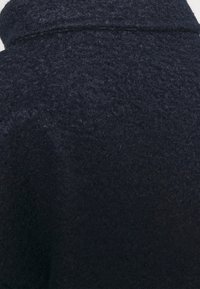 Námořnický modrý vlněný kabát s texturovaným, chlupatým povrchem a vysokým límcem, který se vyznačuje čistým designem bez viditelných zapínání nebo vzorů.