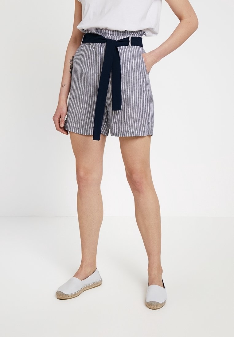 Femme portant un short à rayures verticales grises et blanches avec une ceinture noire, un haut blanc et des chaussures blanches sans lacets, debout, une main dans la poche.