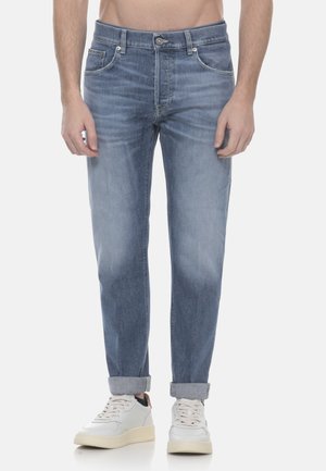 Uomo con jeans azzurri chiari risvoltati e scarpe da ginnastica bianche, in piedi davanti a uno sfondo semplice.